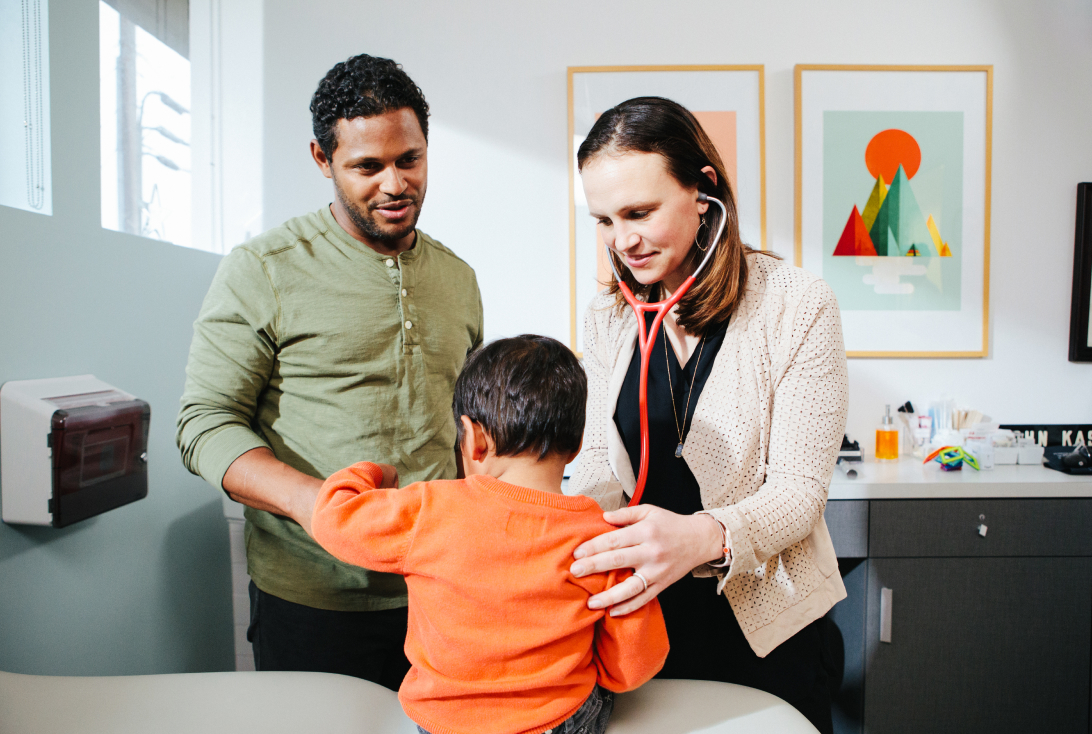 A doctor listening to a young child's chest while his father stands with them